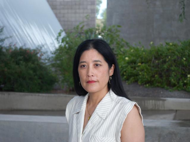 Victoria Chang wears a white shirt and poses for a photograph in front of a garden with grey walls in the background.