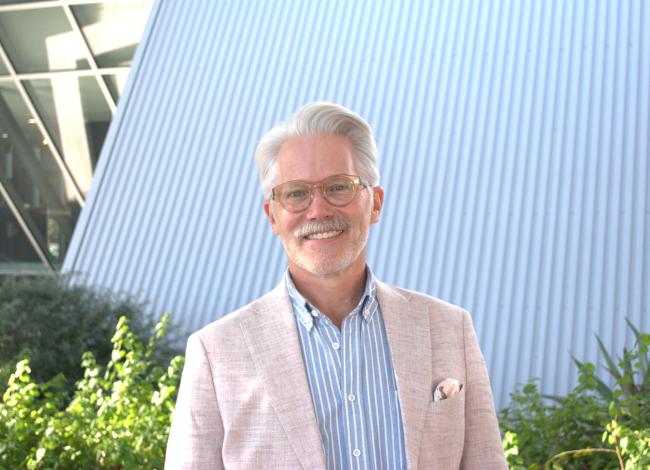 Mark Wunderlich wears a tan blazer over a pale blue button-down with vertical white stripes, as well as glasses with tan, translucent frames. He stands in front of a white, sloped roof with ridges that is next to a glass wall, also sloped. There are also shrubs visible in the background of the photo.