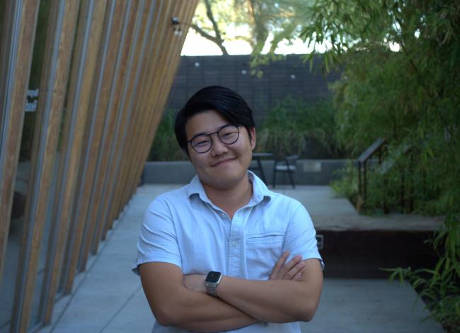 Yanyi stands, smiling, with arms crossed in the Poetry Center's back garden that consists of bamboo and a rock slab. The Poetry Center's slightly curved back wall made of glass and wooden slabs frames the left side of the photo.